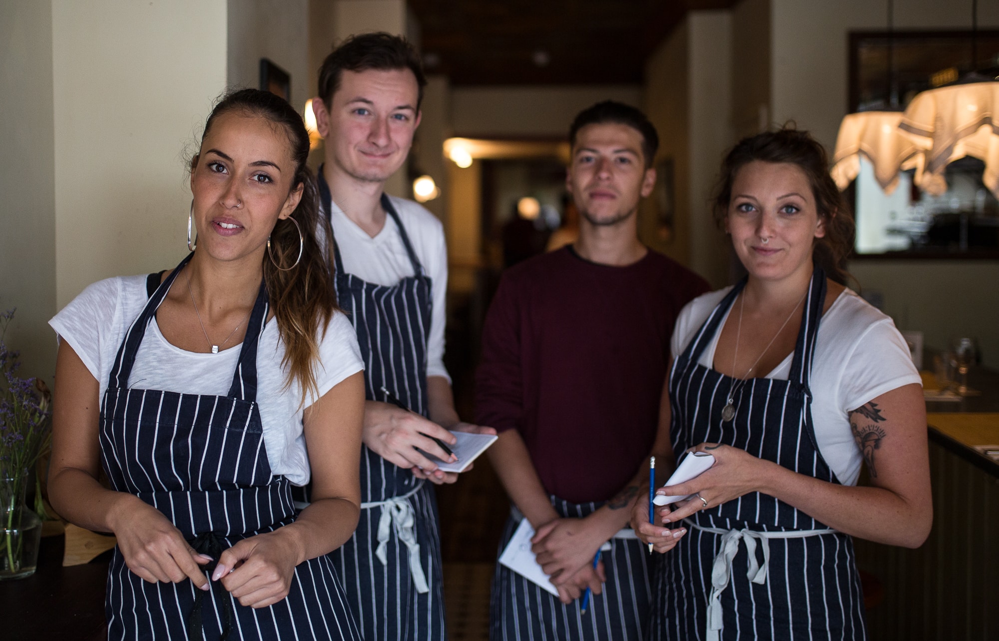 a group of staff working in the hospitality industry in Brighton. Four front of house waiters and waitresses, barman, getting ready for service in a brighton restaurant wearing blue stripy aprons. This is one of the headlining images for Restaurants brighton Jobs