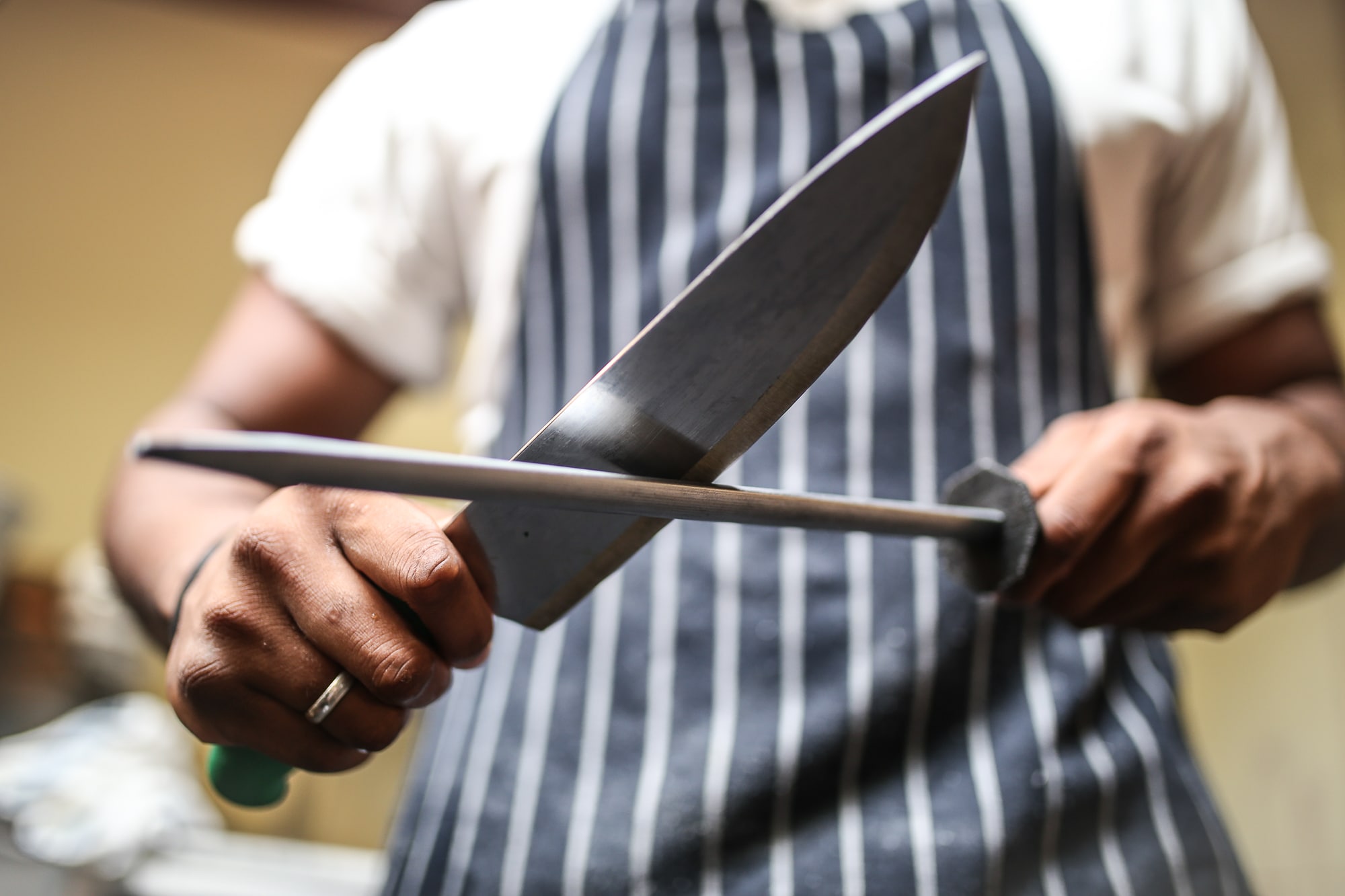 A Brighton chef sharpening his knife at a restaurant in Brighton. The chef in his chef whites with a blue stripey apron. It is a close up of the knife. A headlining image for Restaurants Brighton Jobs