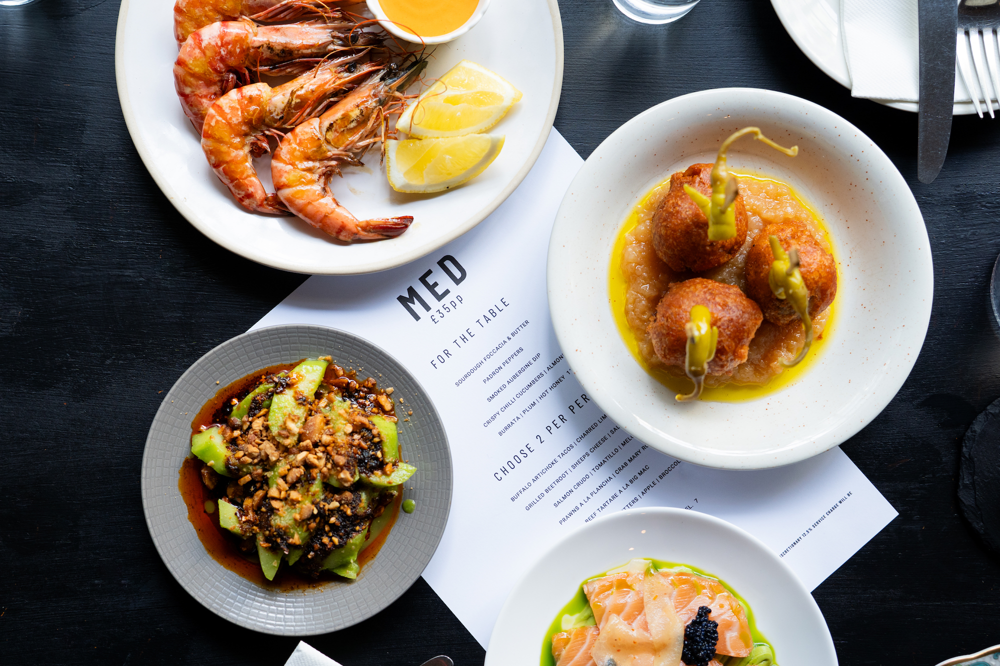 A sharing selection of dishes at Med restaurant in Brighton. Including giant prawns, cured salmon and other colourful dishes. Shot from above on black table. menu is featured. 