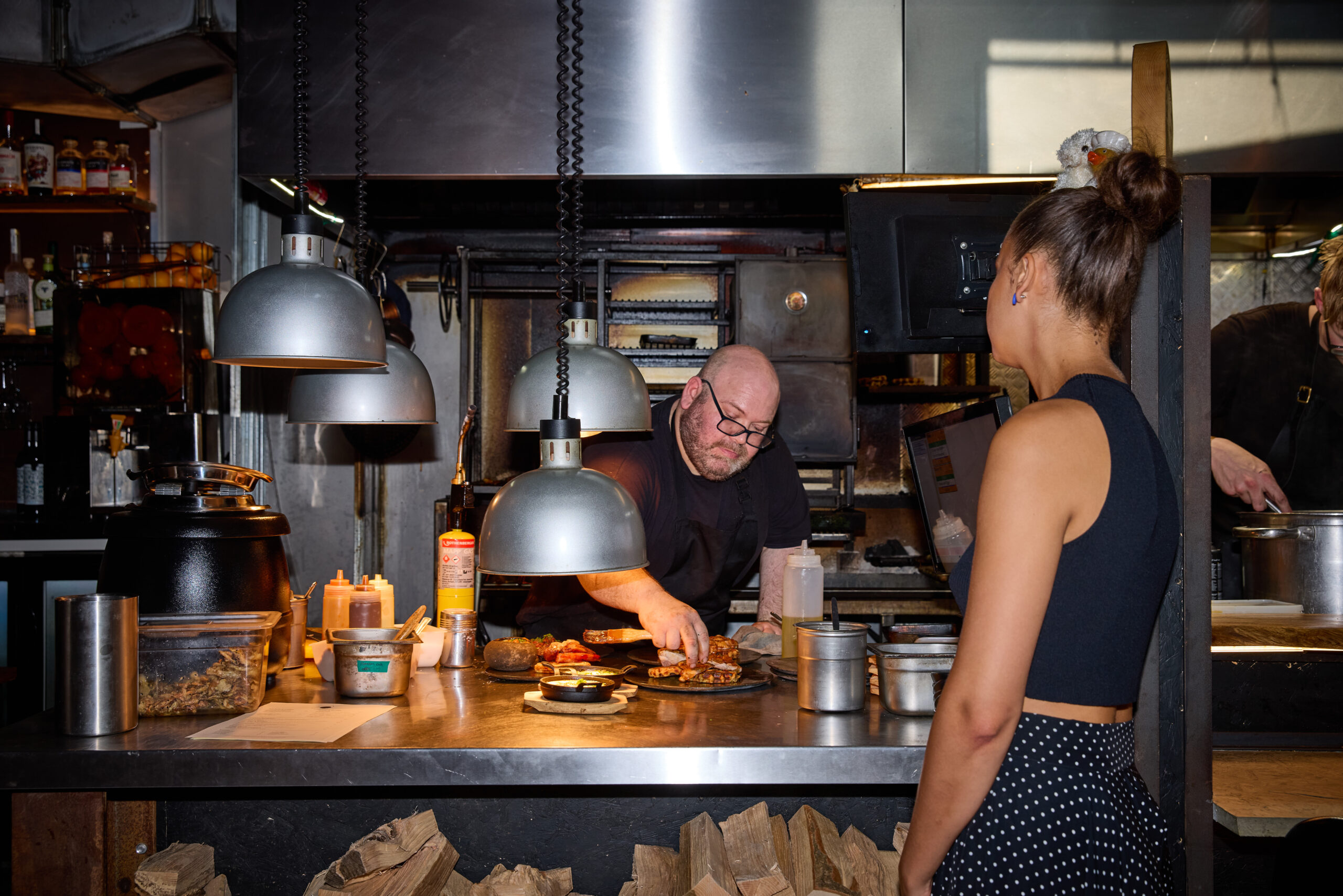 Dave Marrow in his industrial looking kitchen in Brighton's Lanes. A waitress in teh foreground waiting for food. 