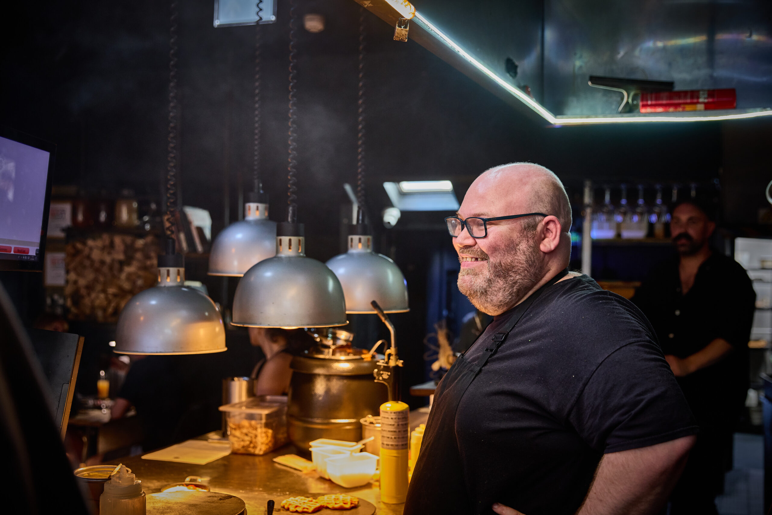 Dave Marrow, the head chef from embers smiling. Stood next to their hot plates at the pass in their open industrial looking kitchen.
