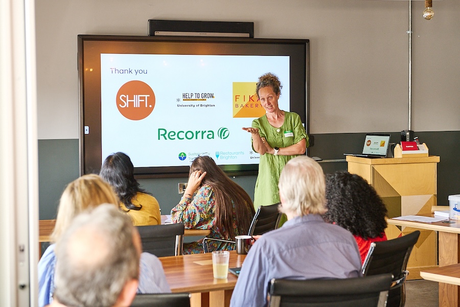 Woman in a green dress standing in front of a screen with logos for Shift, Recorra, Help to Grow and Fika Bakery. She is gesticulating towards the audience. 