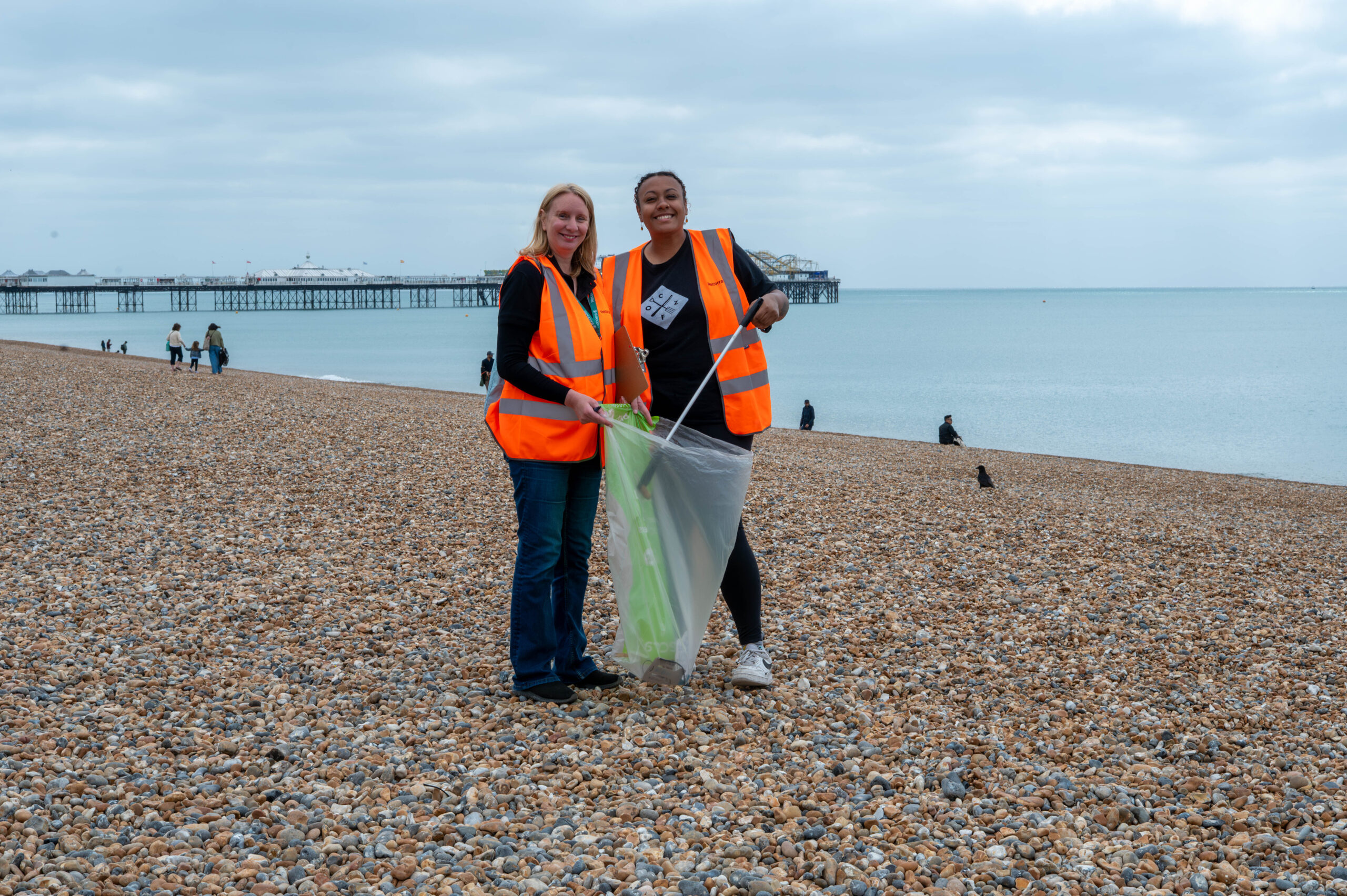 two ladies in orange west on Brighton beach holding bags and collecting plastic waste