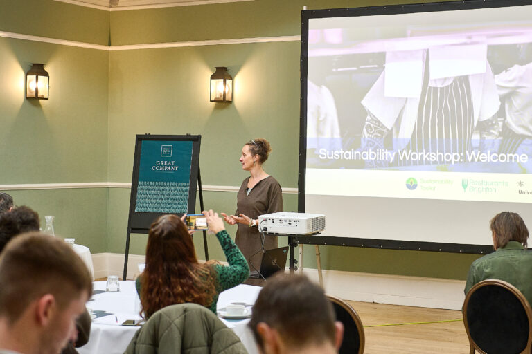 Woman standing by a flip chart in front of a screen with the words sustainability workshop. The slides also show images of chefs.