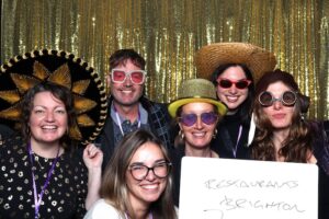 Big smiling faces dressed in silly glasses and hats in front of a gold backdrop holding a white board that says Restaurants Brighton