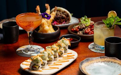 Ellen Richardson – Restaurants Brighton – Blossoms 10.9.25-27 A selection of plates and bowls of sushi and Japanese snacks with cocktails all laid out on a brown wooden table.