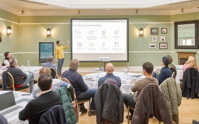 A white man with dark hair wearing a yellow shirt and blue jeans is standing in front or a large projector screen. People are sitting at round tables watching
