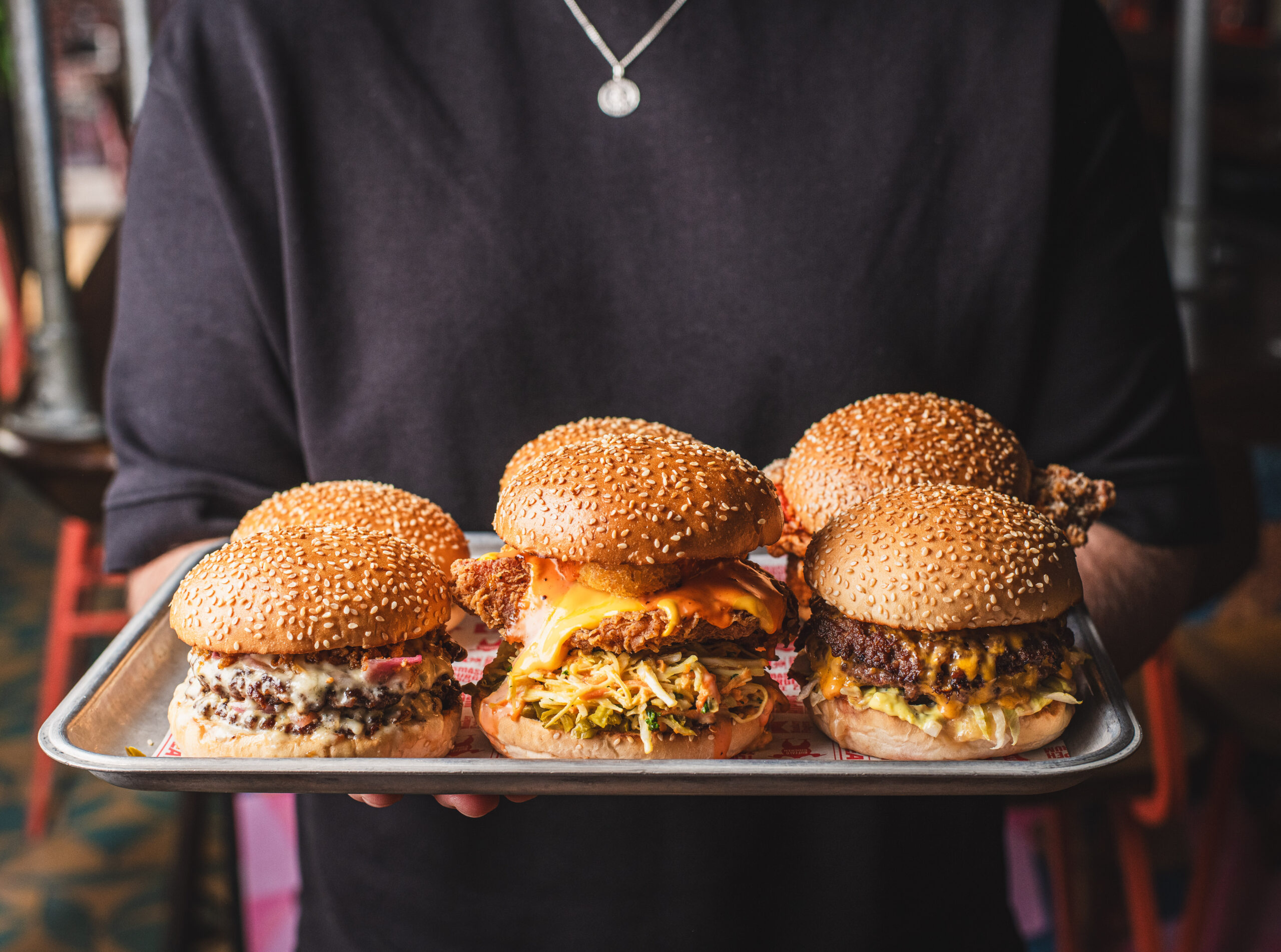 A person holding two trays of food. The trays are laden with burgers, chips, onion rings and lots of indulgent looking food
