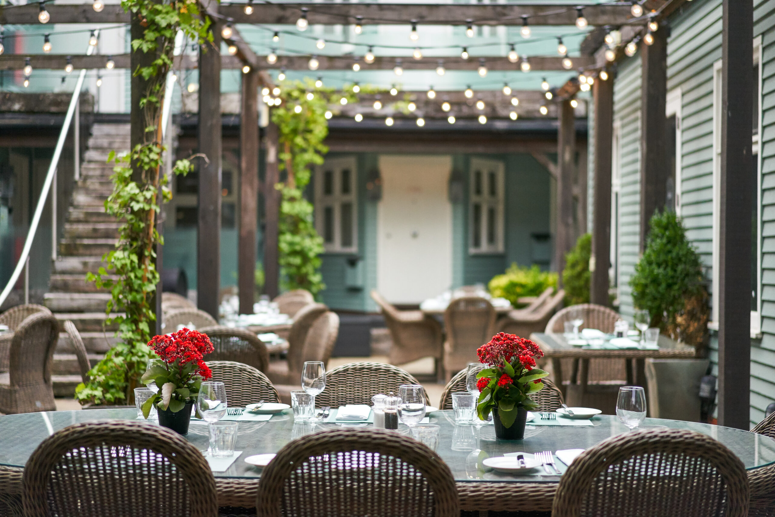 Sage green outdoor area with woonden pagoda, fairy lights, wicker seating and tables dressed with glasses and red flowers.