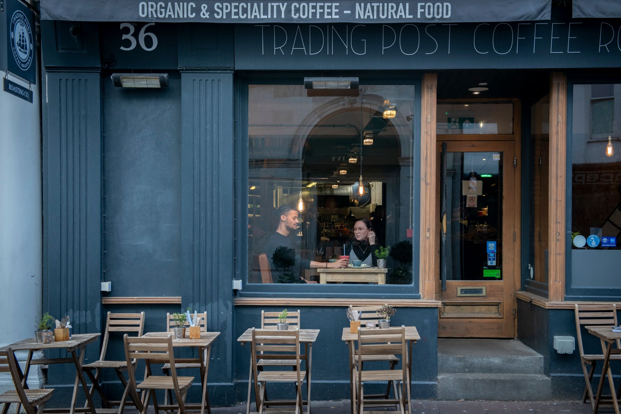 People sitting in the window drinking coffee with alfresco seating in front of them.