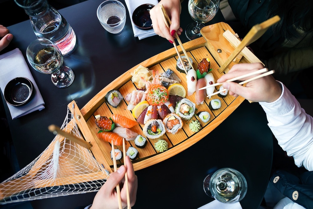Sushi feast served on a wooden boat platter with people using chopsticks to pick pieces of sushi.