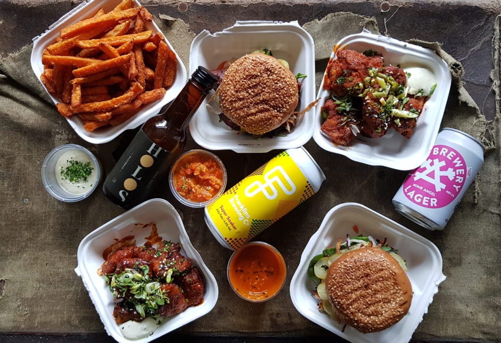 Takeaway boxes of sweet potato fries, cauliflower wins, burgers, dipping sauces and cans of beer.