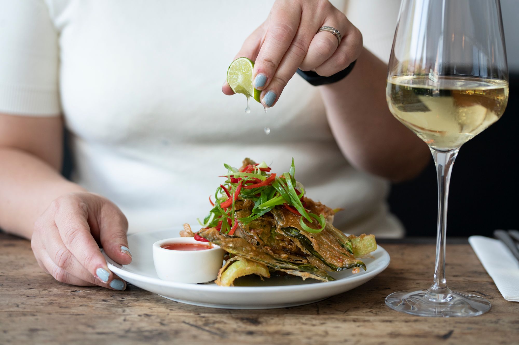 Hand squeezing lime over a plat of colourful food. A glass of white win is at the side of the plate