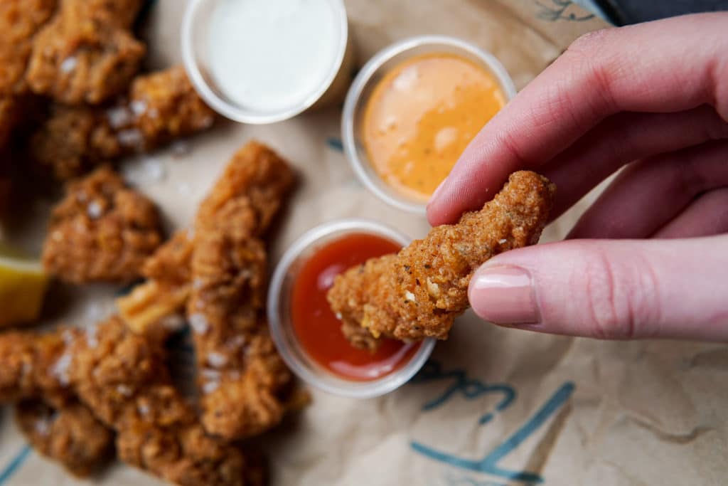 A battered bite being dipped into a pot of sauce, two other with two other sauce pots and more bites laid out on a sheet of food wrapping paper