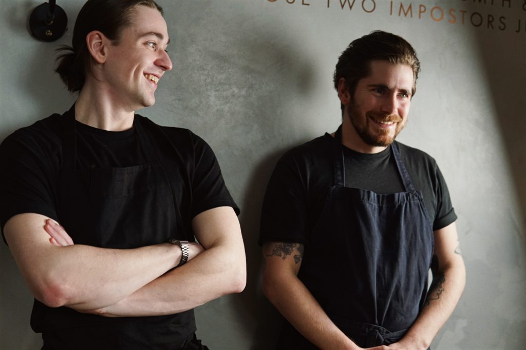 The Tide team, Chefs James and Raph. Raf to the left is earing a balck t-shirt and is looking over his left sholder at James. James, also in a black t-shirt and navy apron holds his hands in his lap and smiles while looking forward.