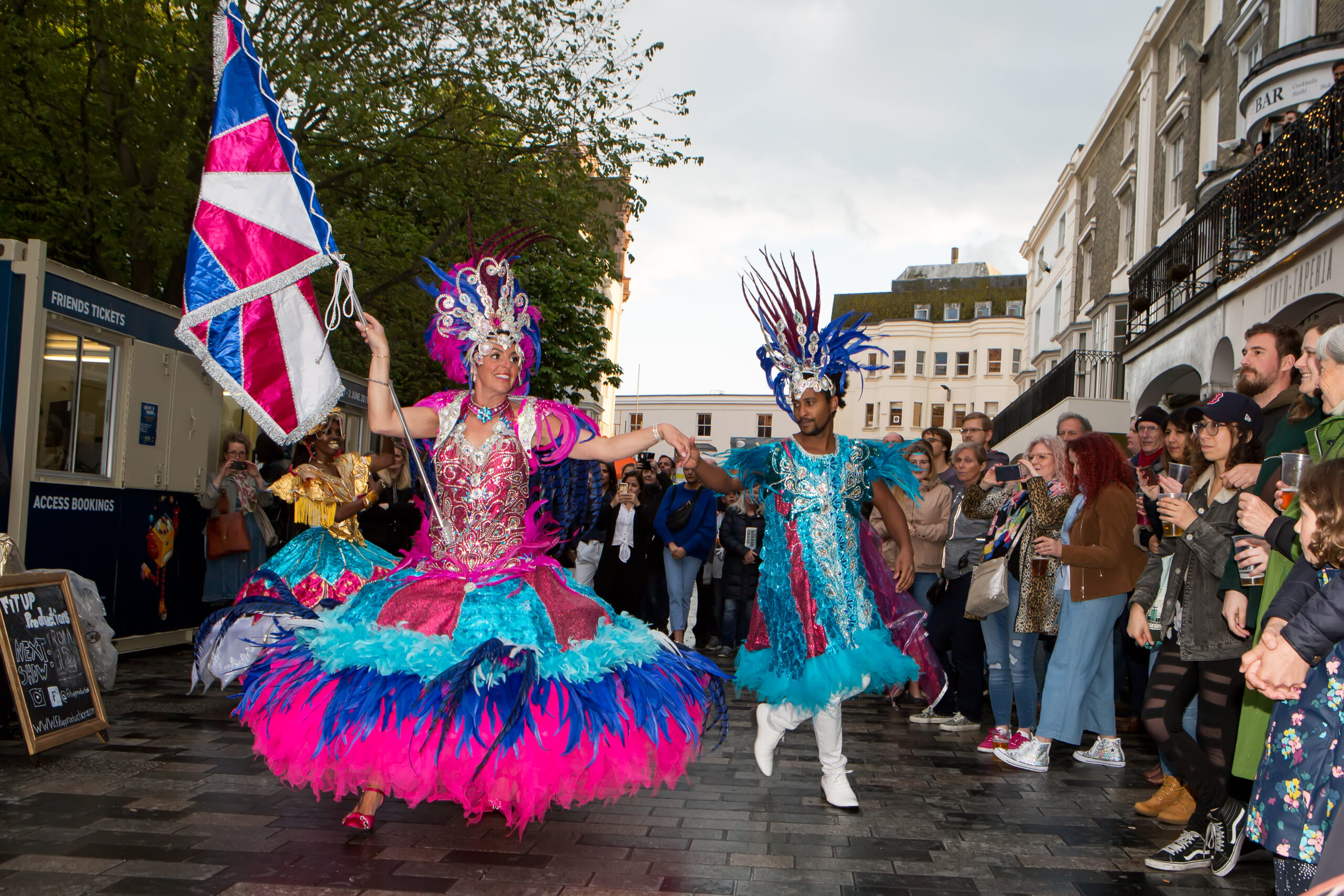 A woman is wearing a wide carnival dress in hot pink and electric blue, she has a feathered silver headdress and carries a pink and blue satine union flag in her right hand.She's holding hands with a man to her left who is wearing an electric blue carnival coat and feathered headdress. The couple are dancing though a street with crowds of onlookers about them. Festivals in Brighton