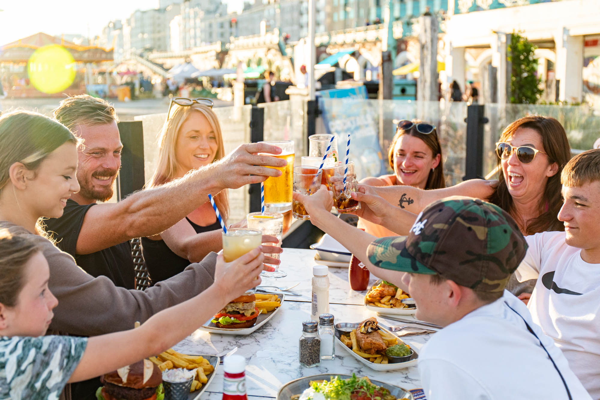 A group of people raising their glasses in a gesture of cheers. They are sitting round a table at a restaurant, outside and the light is warm and bright