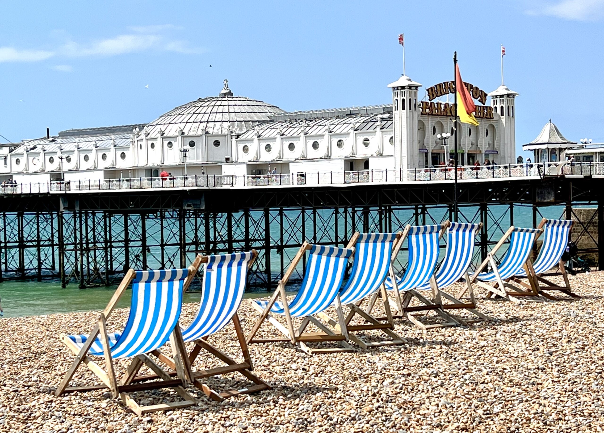 Deck chairs on the pebbles next to Brighton pier on a sunny day with a clear blue sky.