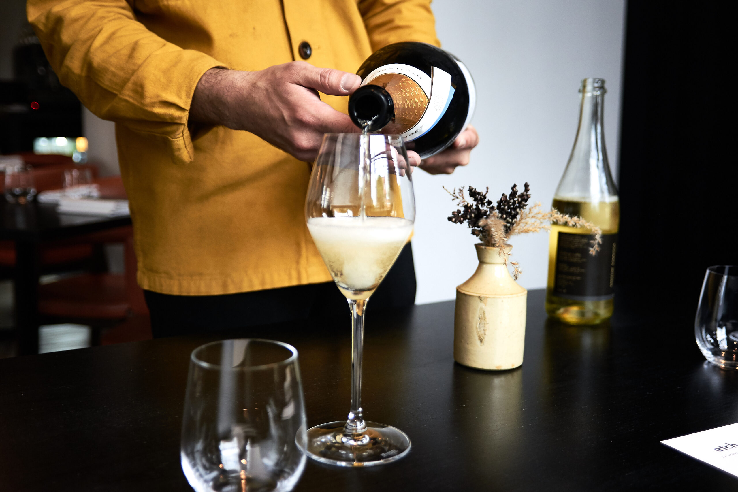 a person in a mustard yellow jacket pouring from a bottle of sparkling wine