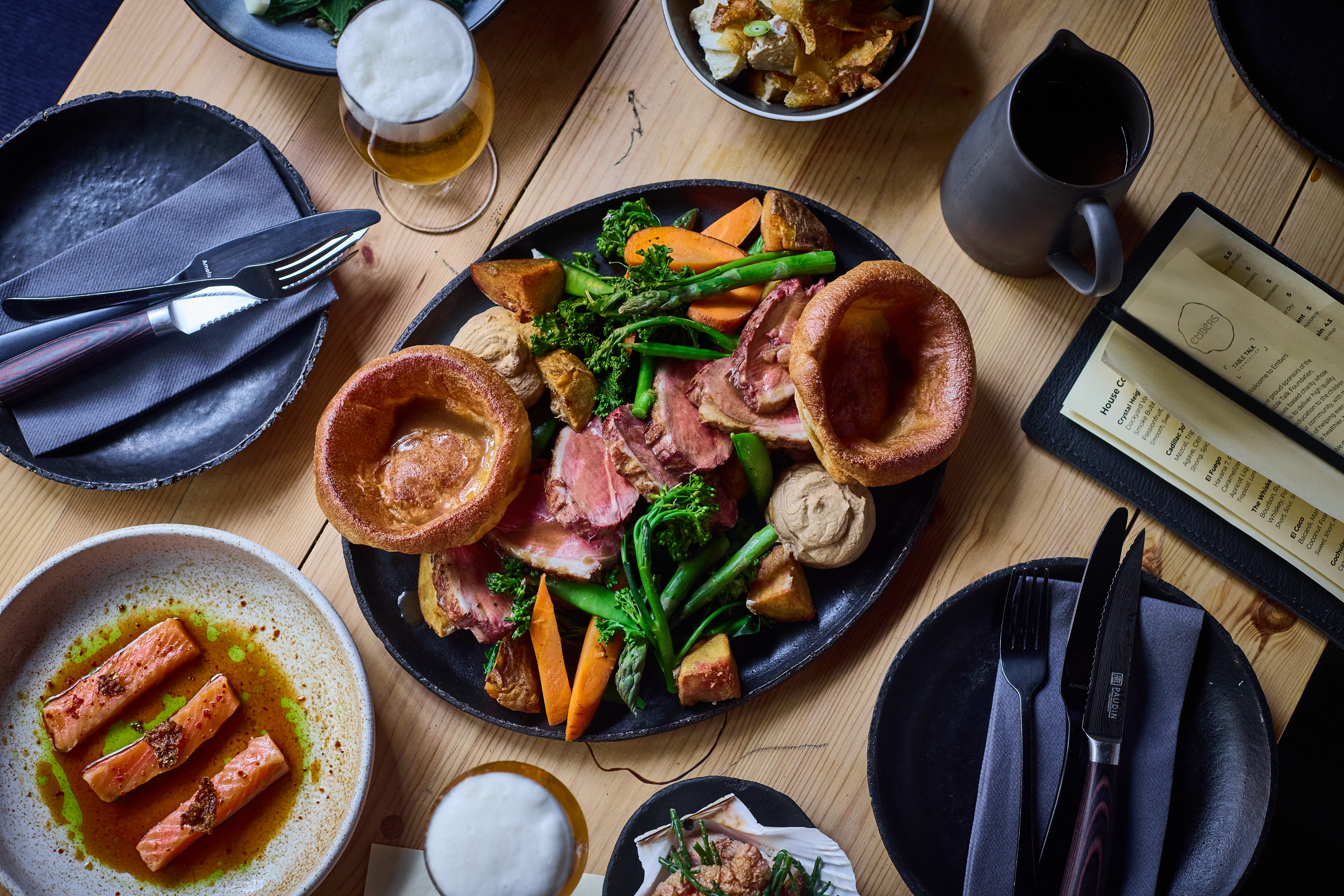 A table photographed from above showing what looks like a sharing platter of a roast. We see carrot, broccoli and yorkshire puddings. The table is set for 2.