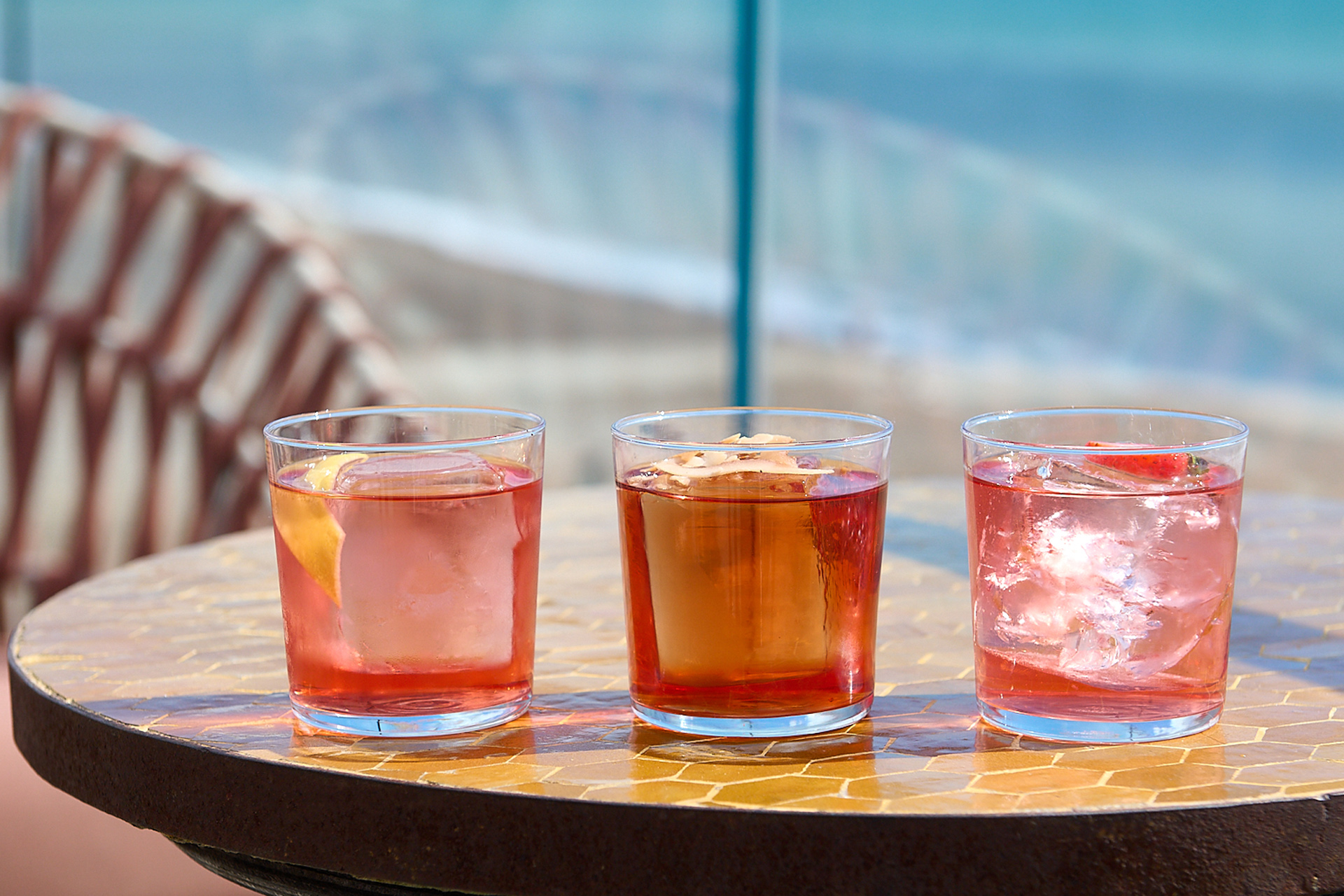 Three short glasses filled with pink looking liquid and ice. Blue sea in the background