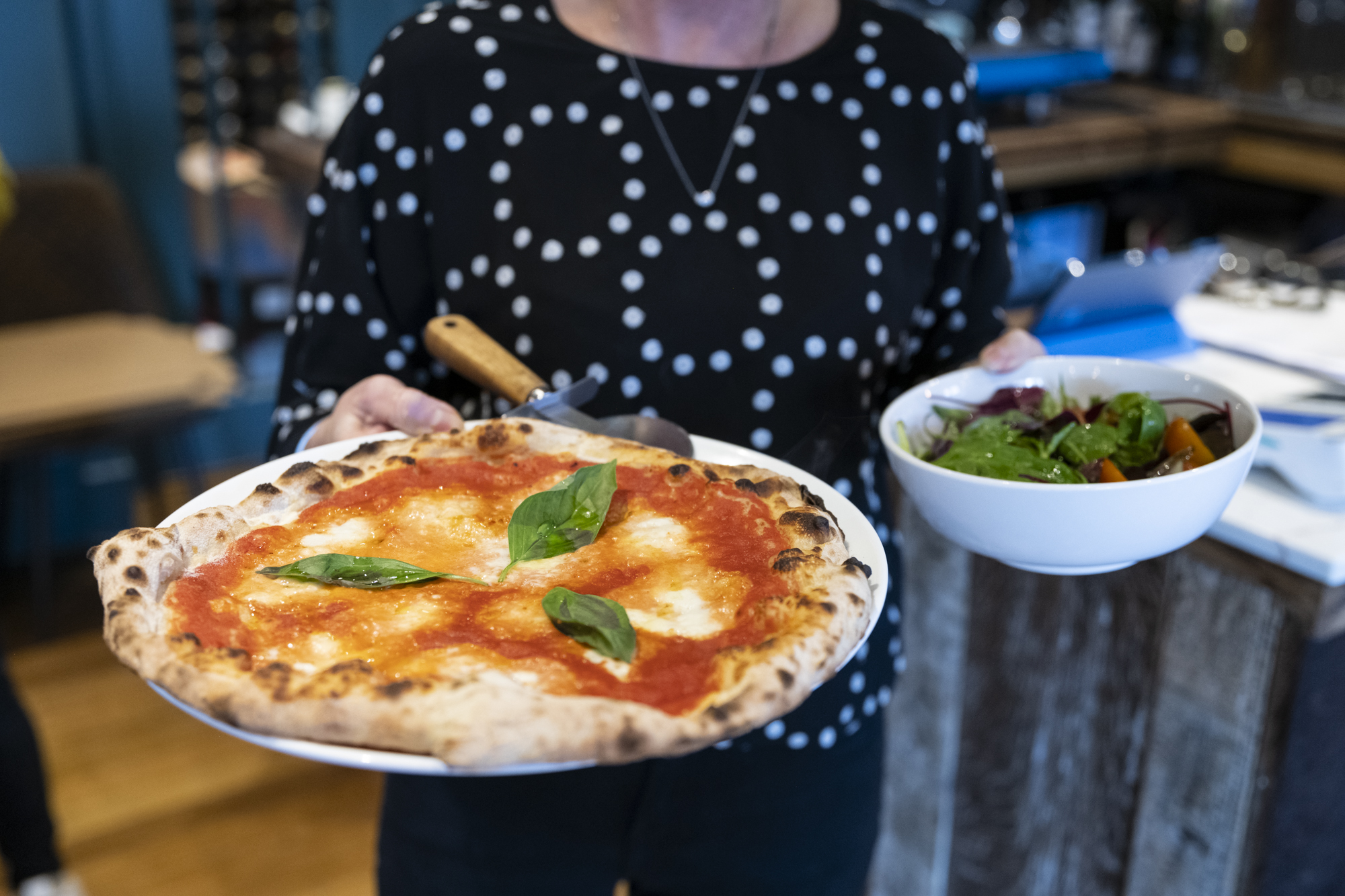 person in a black top with white dotted circles holding a pizza on a plate and a bowl of salad
