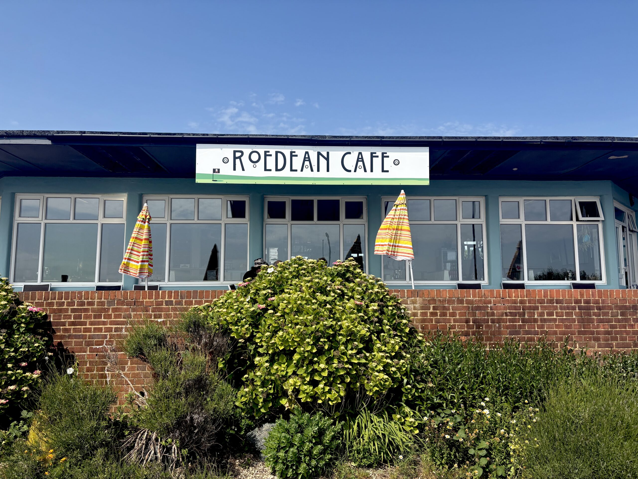 An exterior photo of a cafe against a blue sky. In the foreground is shrubbery and table umbrellas. The cafe has big windows and a sign in art deco lettering that says Roedean Cafe