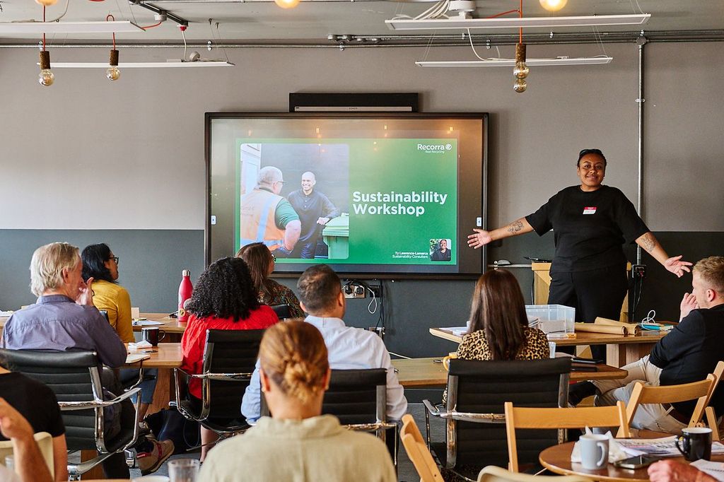 Woman standing with open arms in front of a large screen with the text sustainability workshop. People are watching and listening