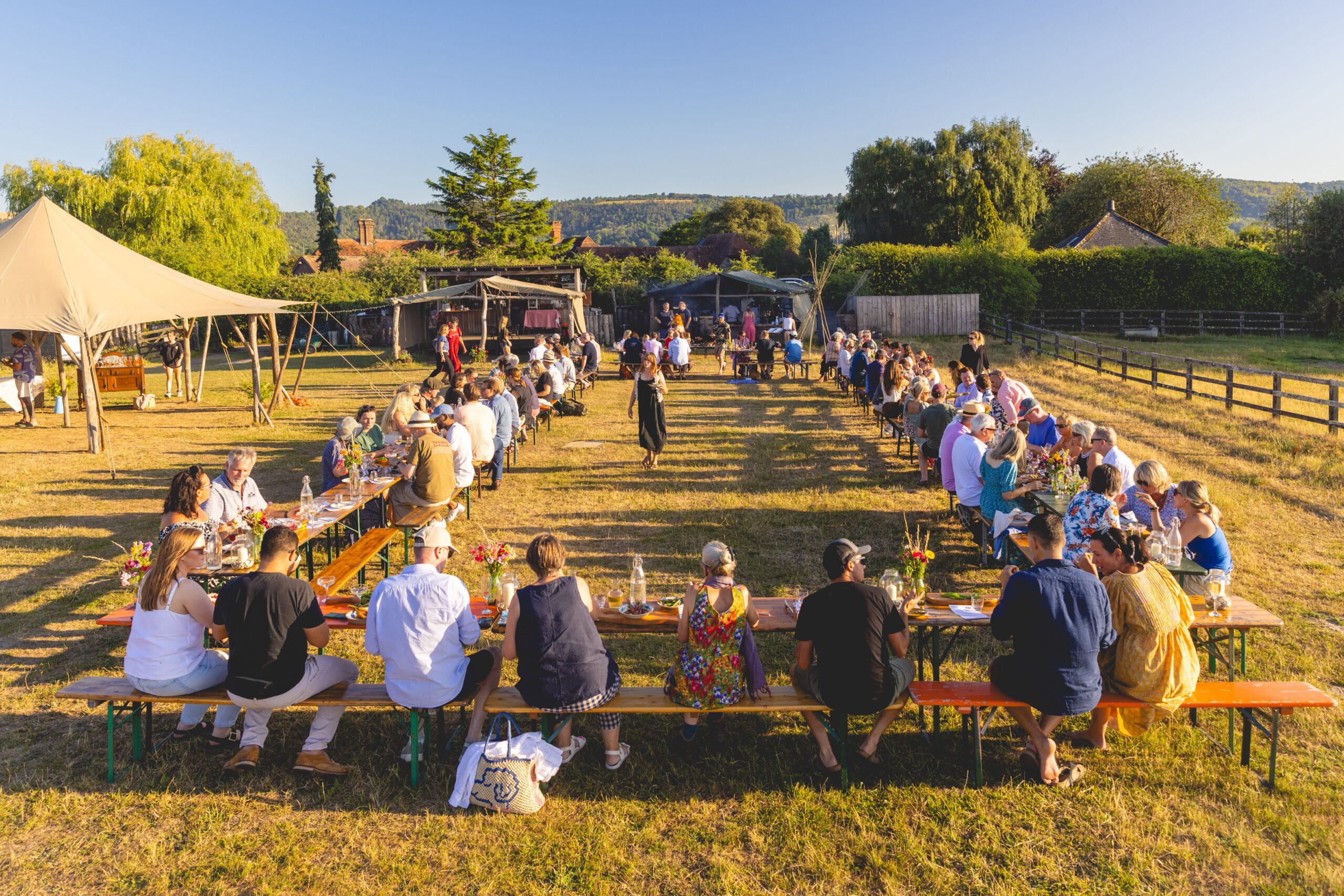 Many people sitting our a large rectangle layout of tables. They are in a field with trees in the background and a canvas shelter. The sun is low and glorious.