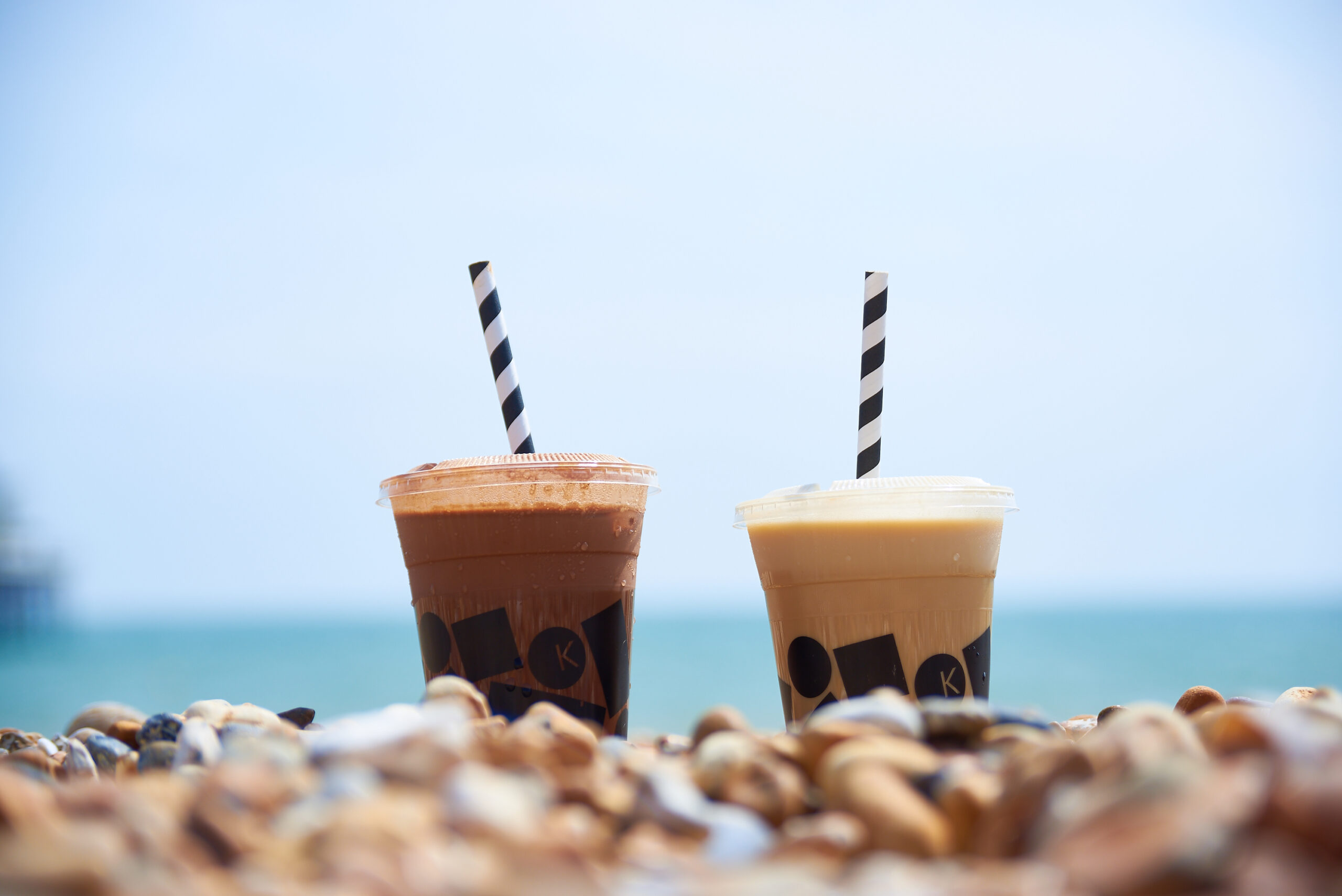 Two plastic cups with blue and white stripy straws sticking out of the top on the beach with the sea in the background. The brown colours indicate one is hot chocolate and the other probably a coffee