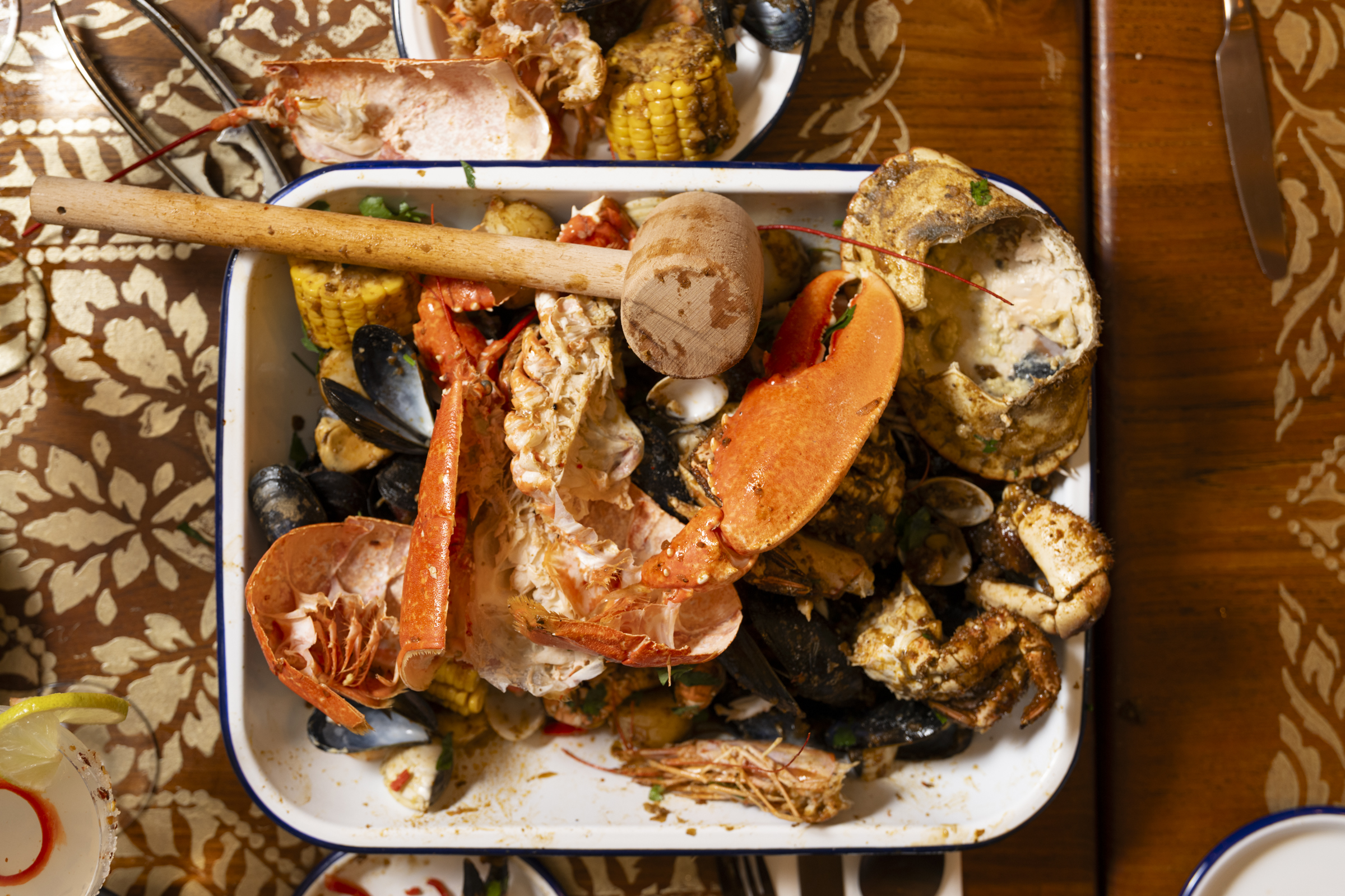 a white enamel tray with blue trim on a brown table. We see remnants of lobster and crab and a wooden hammer