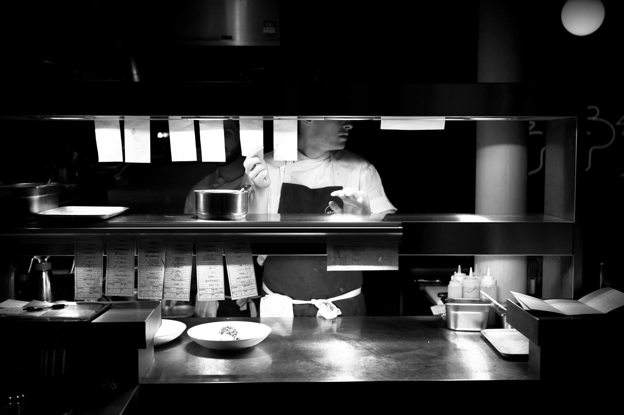 Black and white picture of a chef at the pass. The image is dark, we only see part of the person and we see lots of white bit of paper hanging from the shelves