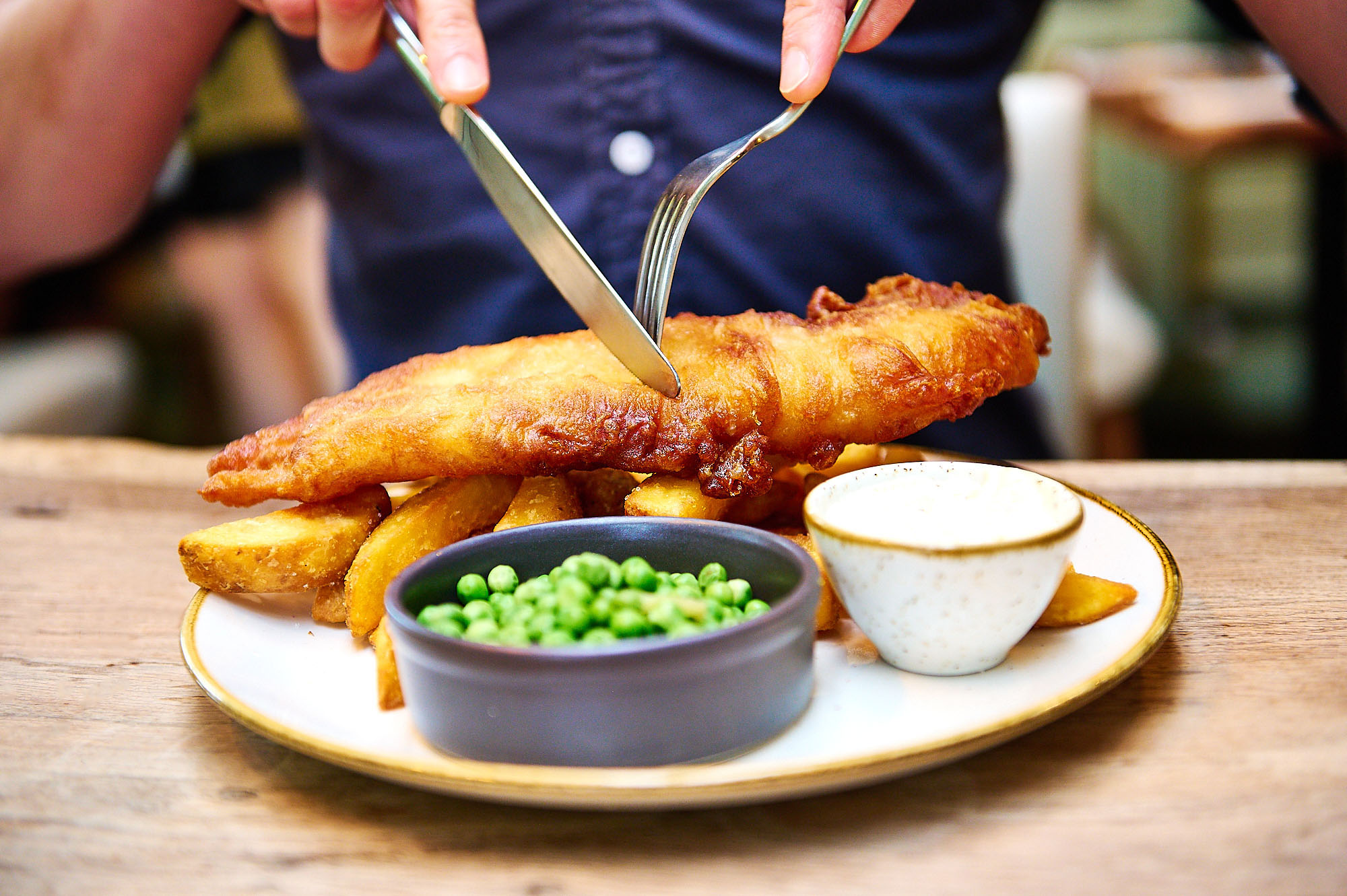 knife and fork about to slice into a crispy looking battered fish on top of chips. Bright green peas in the foreground