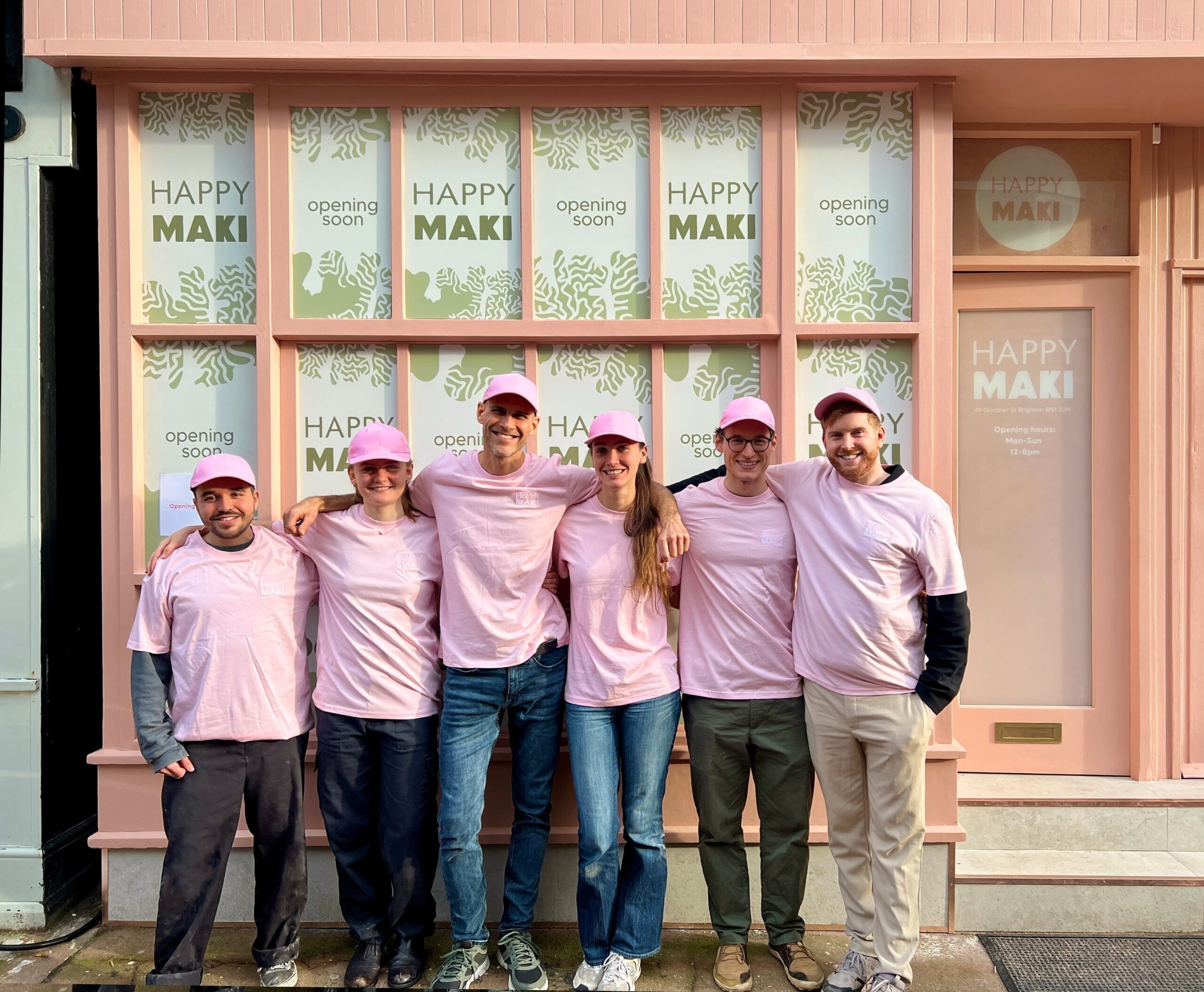 six people standing with their arms around each others shoulders in front of happy maki. they are wearing matching t-shirts and caps
