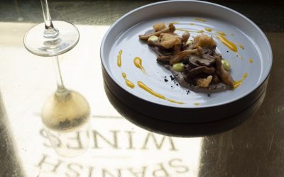 Tapas dish with a dressing on a grey ceramic plate alongside a glass of wine. Photographed in front of the window.